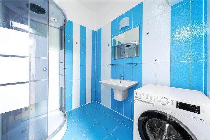 Moderní koupelna s modrou dlažbou, sprchovým koutem a pračkou. Světlý prostor a elegantní provedení vytvářejí příjemnou atmosféru.