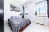 Světlá ložnice s moderním designem, světlou laminátovou podlahou, velkými okny a minimalistickým dekorem.