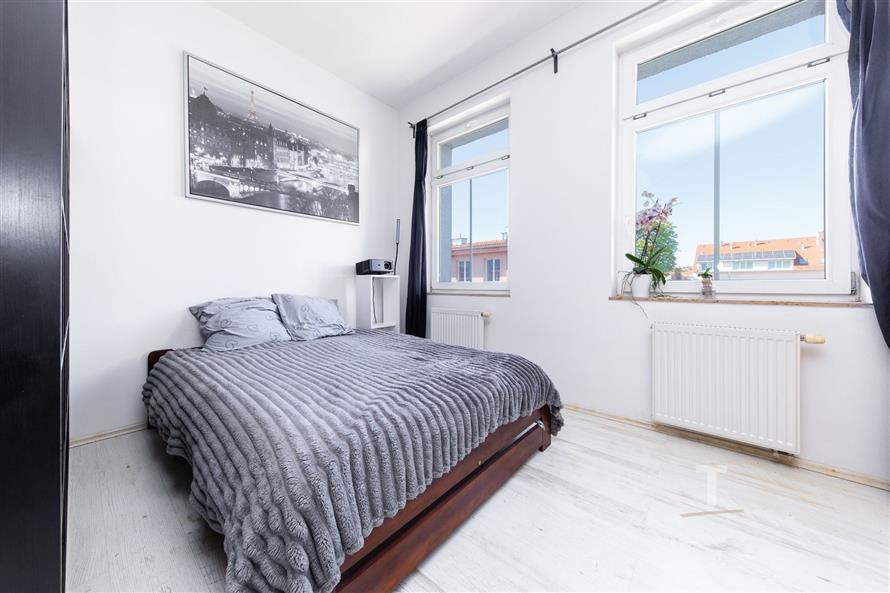 Světlá ložnice s moderním designem, světlou laminátovou podlahou, velkými okny a minimalistickým dekorem.