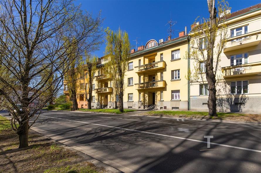 Bytový dům s rekonstruovanými balkony, nové fasády, stromy lemující ulici, slunečný den, klidná rezidenční lokalita.