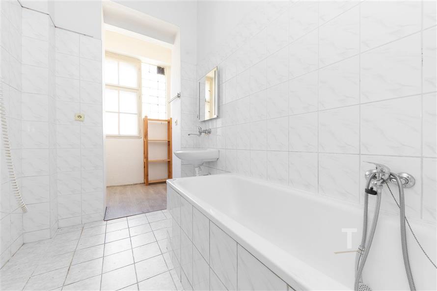 Prostorná koupelna s bílou dlažbou, světlé obklady a velké okno poskytují dostatek přirozeného světla, moderní sanita a jednoduchý design interiéru