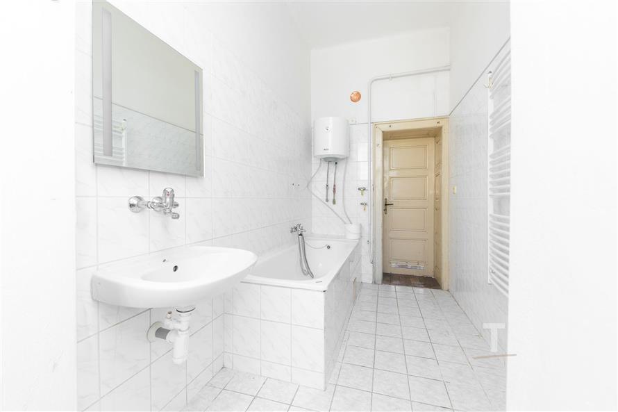Prostorná koupelna s bílými dlaždicemi, vanou a zavěšeným umyvadlem. Světlý interiér, k dispozici žebříkové topení a elektrický bojler.