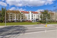 Nízkopodlažní bytový komplex s červenou střechou, moderní fasádou a velkými balkony, obklopený zelení a parkovacími místy na klidné ulici.