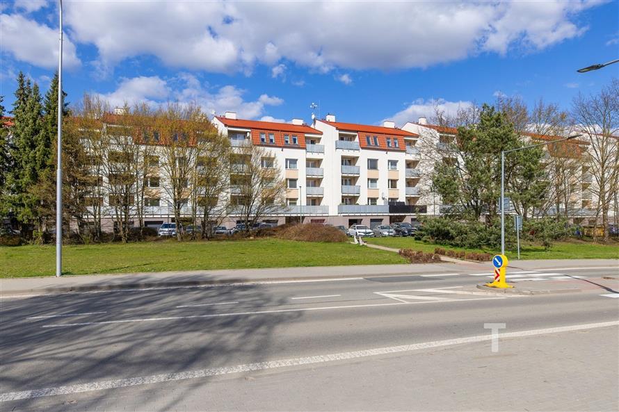 Nízkopodlažní bytový komplex s červenou střechou, moderní fasádou a velkými balkony, obklopený zelení a parkovacími místy na klidné ulici.
