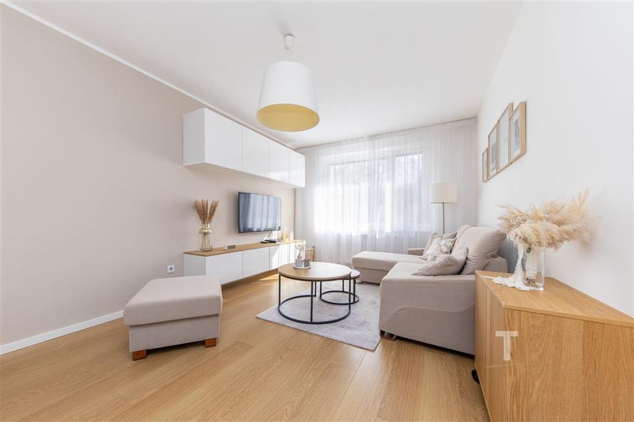 Světlý obývací pokoj s parketovou podlahou, moderní nábytkem a velkými okny, které zajišťují dostatek přirozeného světla.