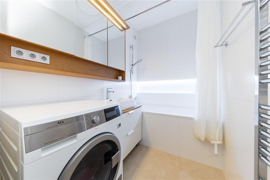 Koupelna s moderní pračkou, světlá dlažba, velké zrcadlo a vana, vše v minimalistickém stylu a s dřevěnými prvky