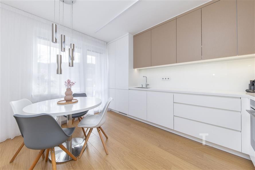 Moderní kuchyň s dřevěnou podlahou, vestavěné skříně a světlým designem, kulatý jídelní stůl s elegantními židlemi u velkého okna