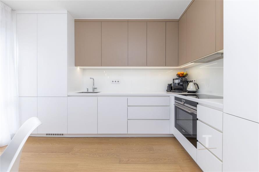 Moderní kuchyň s bílými skříňkami, dřevěnou podlahou a vestavěnými spotřebiči. Světlý prostor s elegantním designem.