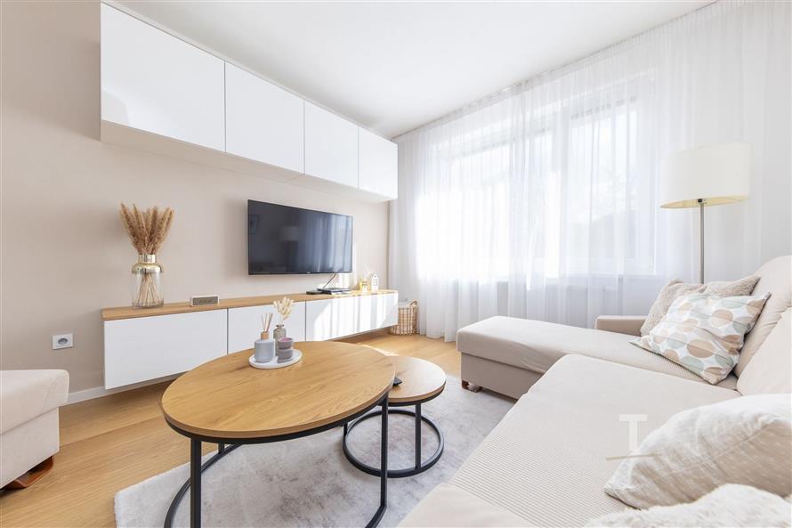 Světlý obývací pokoj s parketovou podlahou, velká okna s bílými závěsy, moderní nábytek a neutrální barevná paleta nabízí útulný prostor.