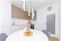 Moderní kuchyň s bílými skříňkami, vestavěnými spotřebiči a světlou dlažbou, doplněná stylovým osvětlením a nástěnnými hodinami.