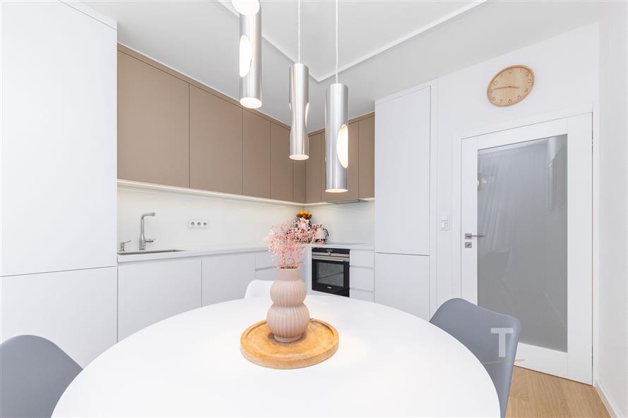 Moderní kuchyň s bílými skříňkami, vestavěnými spotřebiči a světlou dlažbou, doplněná stylovým osvětlením a nástěnnými hodinami.