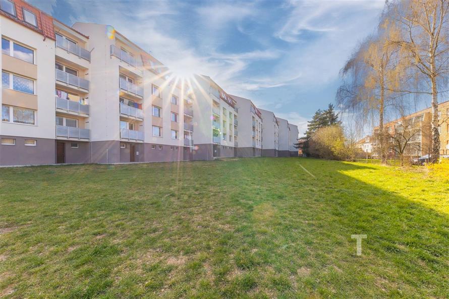 Exteriér bytového domu s udržovaným trávníkem, balkony a moderní fasádou, v klidné rezidenční oblasti.