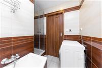 Moderní koupelna s dlažbou a dřevěnými obklady, obsahující vanu a sprchový kout. Pračka umístěná u dveří dodává praktičnost prostoru.