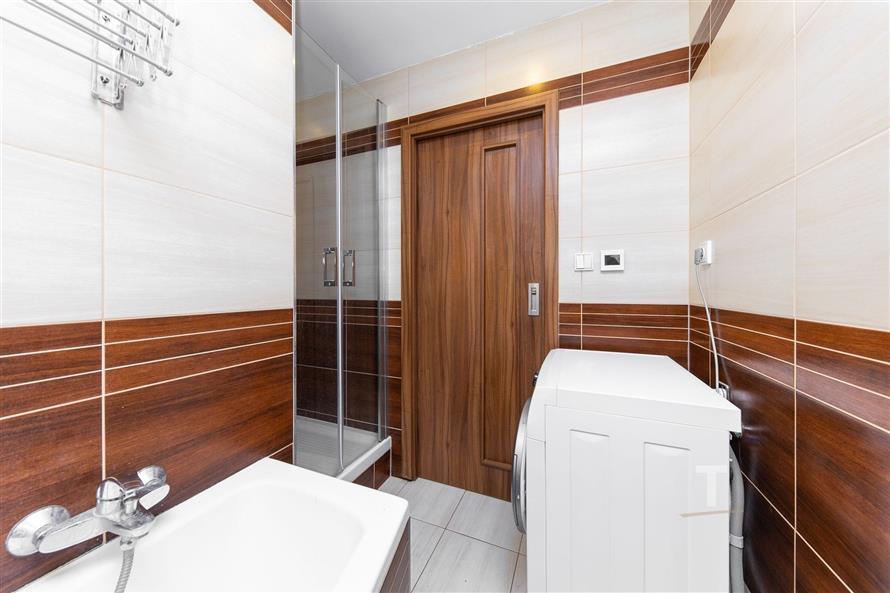 Moderní koupelna s dlažbou a dřevěnými obklady, obsahující vanu a sprchový kout. Pračka umístěná u dveří dodává praktičnost prostoru.