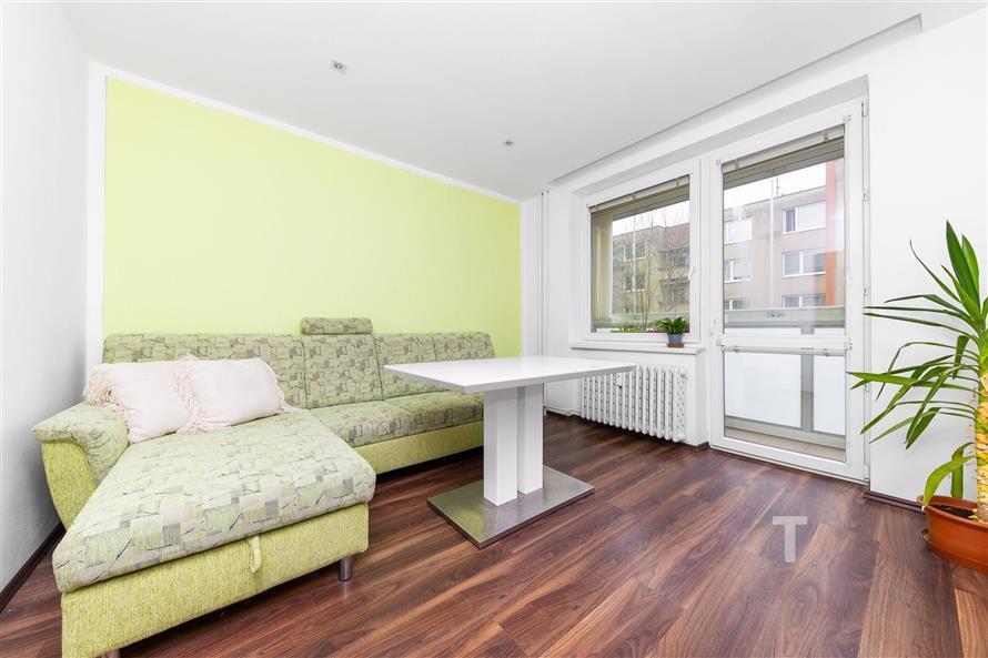 Světlý obývací pokoj s laminátovou podlahou, velká okna s výhledem, moderní pohovka a zelená stěna vytváří útulný prostor.