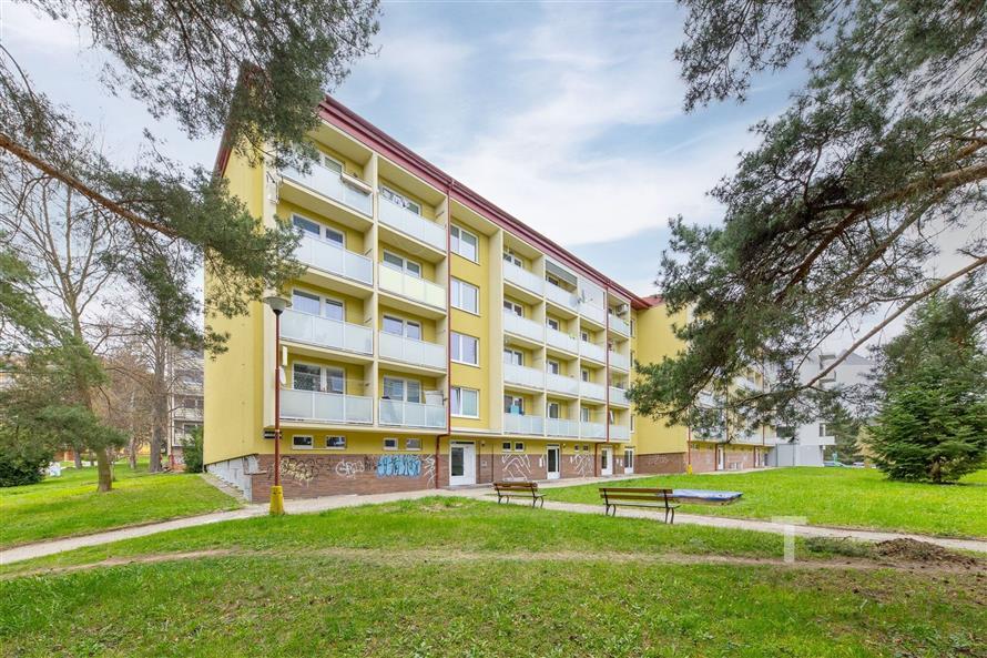 Čtyřpatrový panelový bytový dům s balkony, obklopen zelení, ideální pro klidné bydlení v rezidenční čtvrti, poblíž parků a služeb.