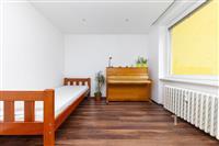 Jednoduchý pokoj s parketovou podlahou, dřevěnou postelí a pianem. Velké okno propouští dostatek světla, bílá stěna vytváří čistý vzhled.