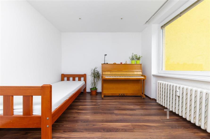 Jednoduchý pokoj s parketovou podlahou, dřevěnou postelí a pianem. Velké okno propouští dostatek světla, bílá stěna vytváří čistý vzhled.