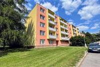 Barevný činžovní dům s balkonem, obklopený zelení a s parkovištěm v blízkosti, vhodný pro rodinné bydlení v klidné lokalitě.