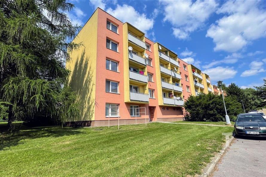 Barevný činžovní dům s balkonem, obklopený zelení a s parkovištěm v blízkosti, vhodný pro rodinné bydlení v klidné lokalitě.