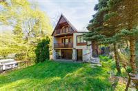 Přední pohled na rekreační chatu v přírodním prostředí, s dřevěnými balkony, obklopená zelení a vzrostlými stromy, nabízí klidnou atmosféru.