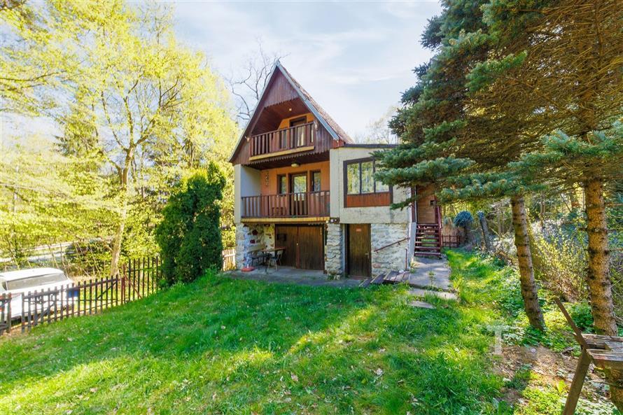 Přední pohled na rekreační chatu v přírodním prostředí, s dřevěnými balkony, obklopená zelení a vzrostlými stromy, nabízí klidnou atmosféru.