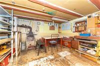 Dílna s dlažbou, pracovní stoly a policové regály. Obsahuje kamna na dřevo a nářadí, ideální pro kutilské projekty či skladování.