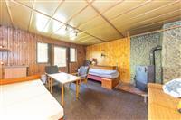 Útulný obývací pokoj s dřevěným obložením, krbovými kamny a velkými okny, které dodávají prostoru světlost.