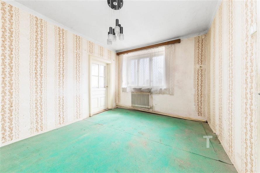 Prázdná místnost s oknem, zeleným kobercem, tapetovanými stěnami a původním radiátorem, ideální pro renovaci nebo modernizaci prostoru.