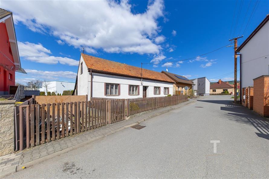 Tradiční venkovská ulice s rodinným domem, červenou střechou a dřevěným plotem, okolní klidné prostředí a slunečné počasí.