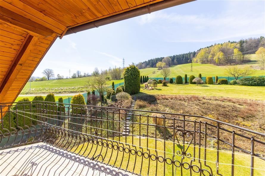 Balkón s výhledem na zahradu, kovové zábradlí a dřevěný strop, okolní krajina se zelenými loukami a kopci, ideální pro klidný život.