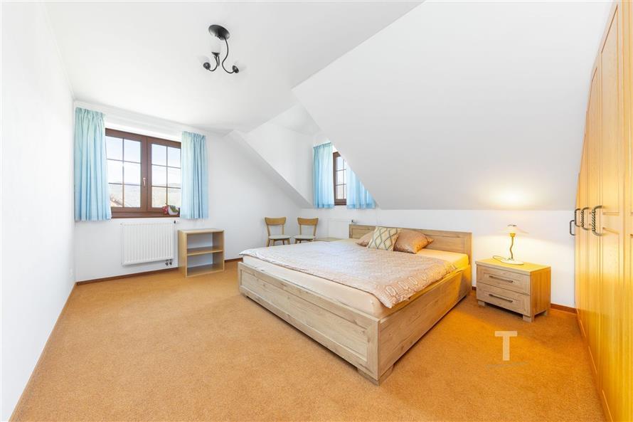 Prostorná ložnice s dřevěnou postelí, velká okna s modrými závěsy, světlý interiér, vestavěné skříně, koberec na podlaze