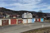 Garážová jednotka s plechovými vraty, nacházející se v klidném venkovském prostředí s výhledem na zalesněné kopce a rodinné domy v pozadí.