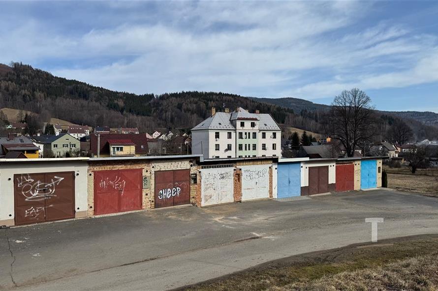 Garážová jednotka s plechovými vraty, nacházející se v klidném venkovském prostředí s výhledem na zalesněné kopce a rodinné domy v pozadí.