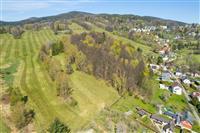 Zasněžená kopcovitá krajina s rozmanitými lesy a loukami, sousedící s malou vesnicí a venkovskými domy, obklopená zelení a přírodními scenériemi.