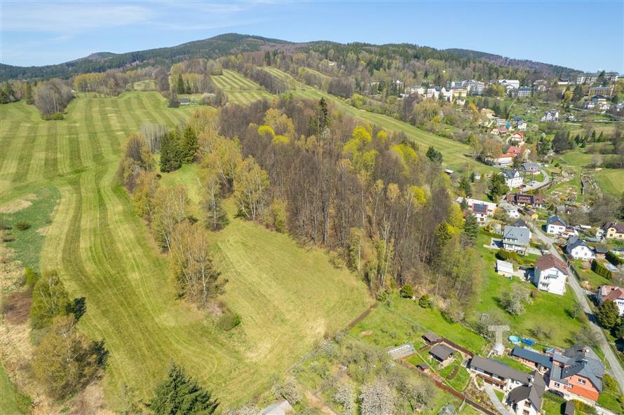 Zasněžená kopcovitá krajina s rozmanitými lesy a loukami, sousedící s malou vesnicí a venkovskými domy, obklopená zelení a přírodními scenériemi.