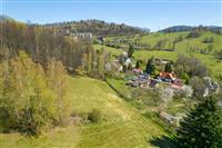 Pohled na malebnou obec obklopenou zelenými loukami a lesy, v pozadí kopce a moderní domy, klidné venkovské prostředí.