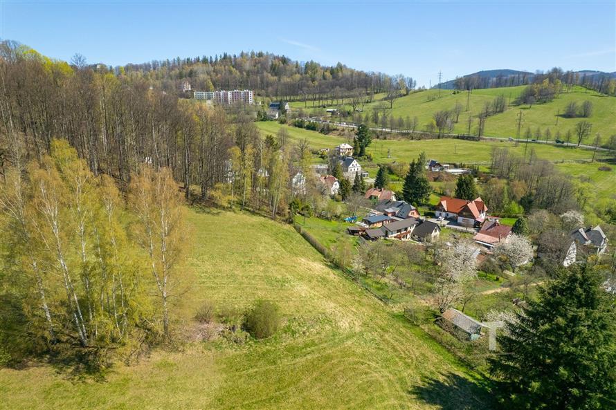 Pohled na malebnou obec obklopenou zelenými loukami a lesy, v pozadí kopce a moderní domy, klidné venkovské prostředí.