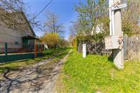 Úzká přístupová cesta lemovaná zelenými ploty vede k venkovskému domu, obklopena stromy a přírodními prvky, vhodná lokalita pro klidné bydlení.