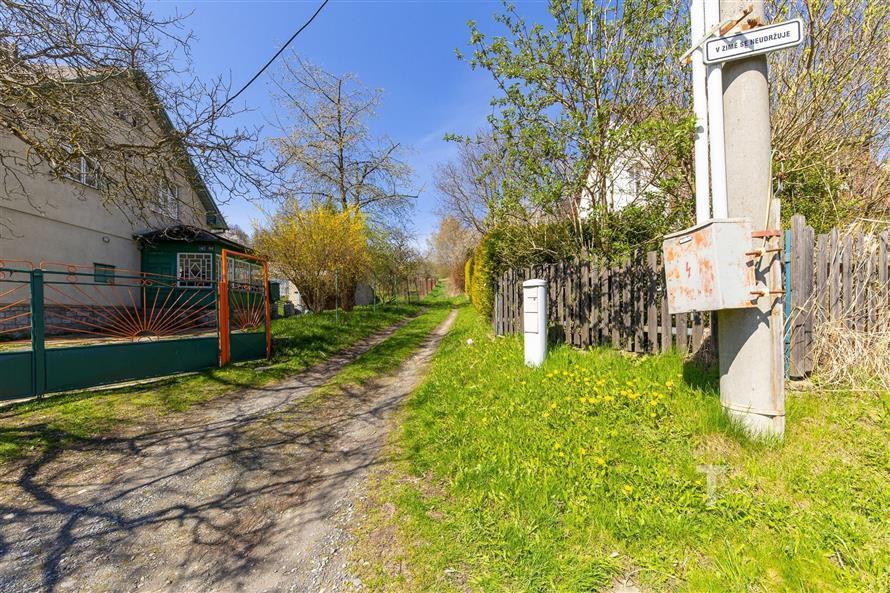 Úzká přístupová cesta lemovaná zelenými ploty vede k venkovskému domu, obklopena stromy a přírodními prvky, vhodná lokalita pro klidné bydlení.